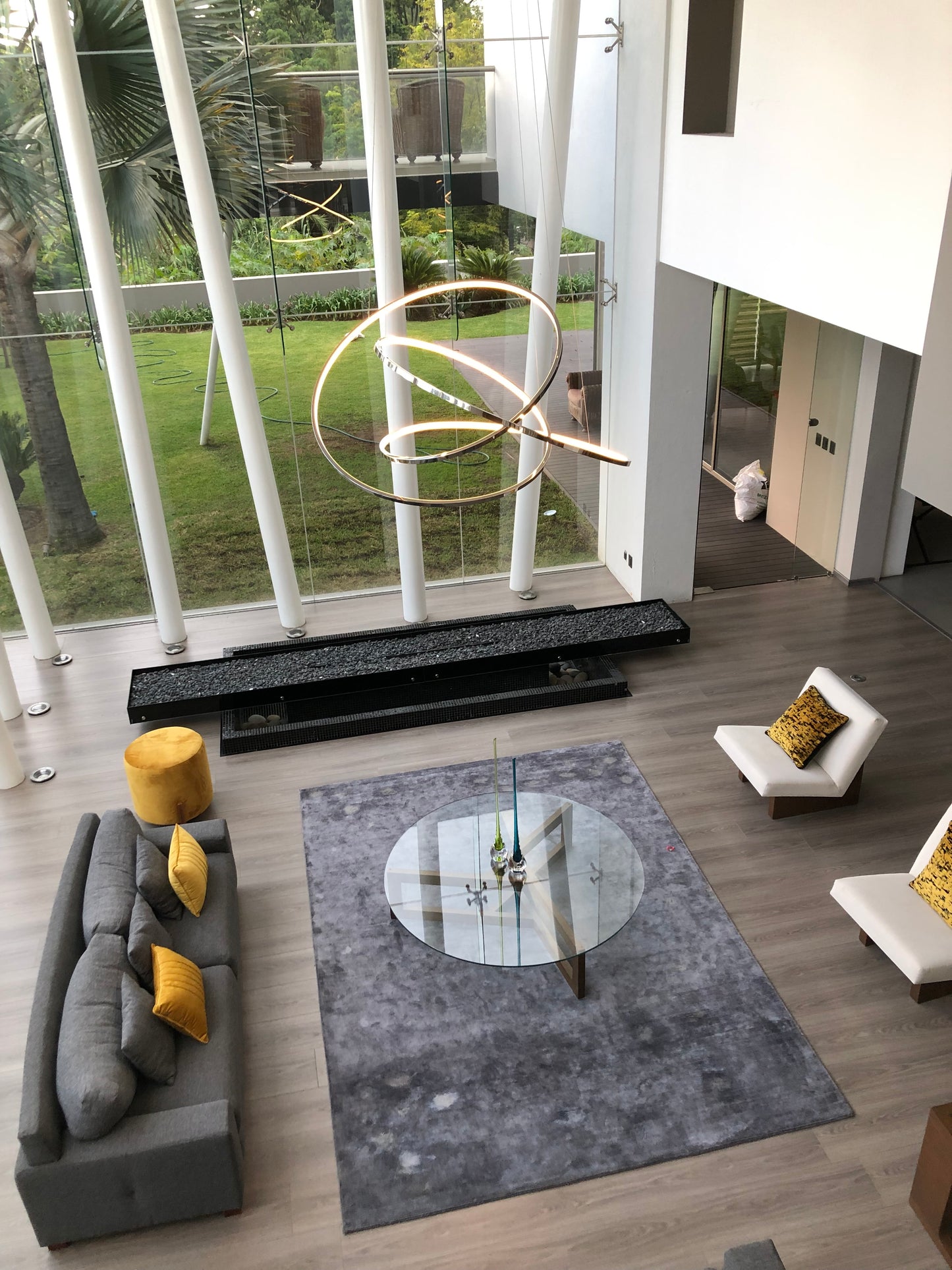 Sala moderna con lámpara colgante dorada, ventanales altos, sofá gris y mesa de cristal.