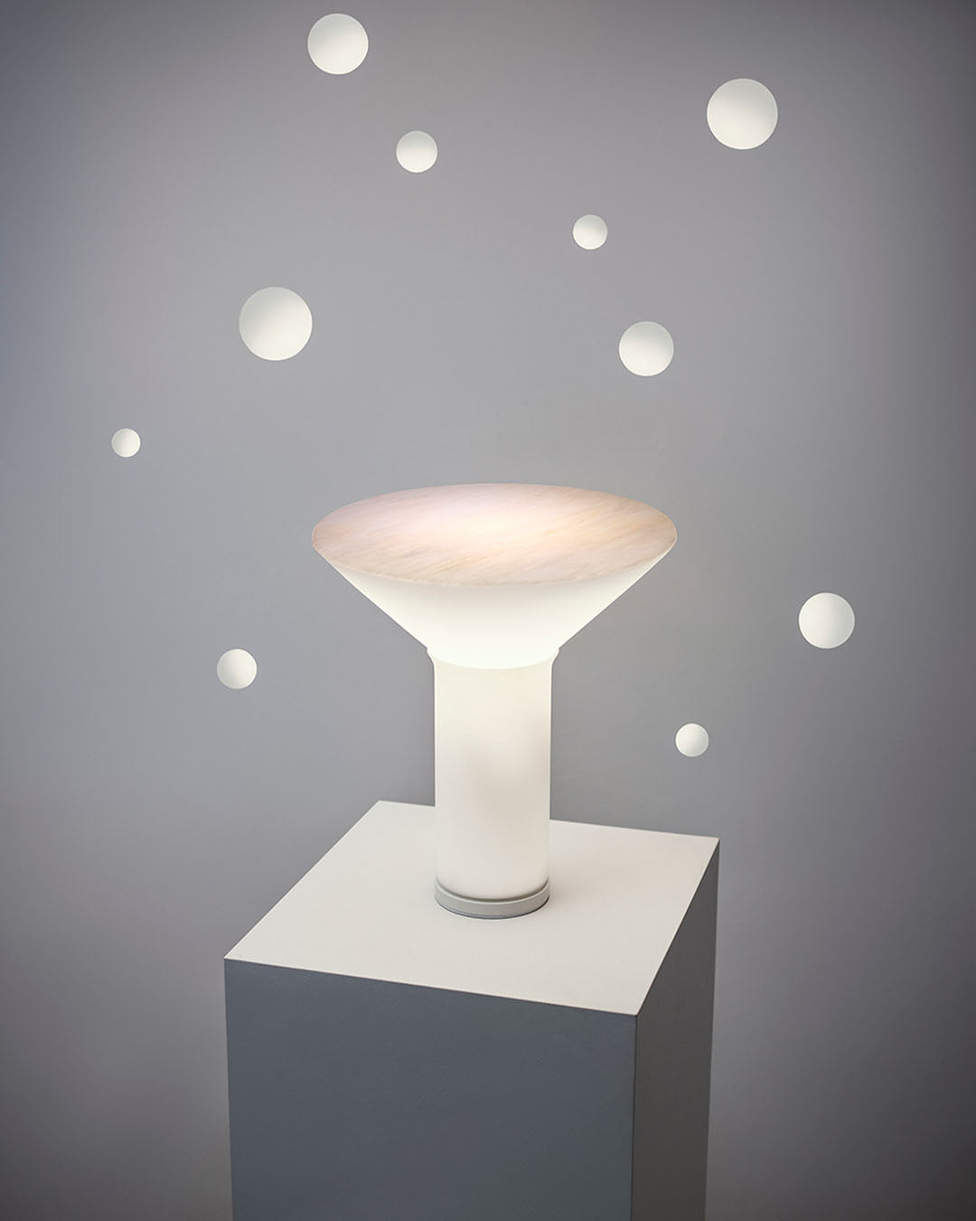 Lámpara de mesa moderna blanca en pedestal, fondo gris con detalles circulares, Birot MX iluminación.