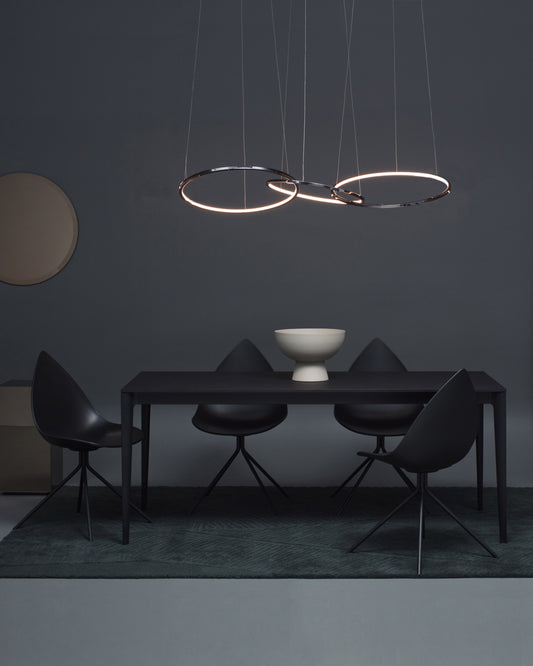 Lámpara colgante moderna de aro LED sobre mesa negra con sillas y decoración minimalista.