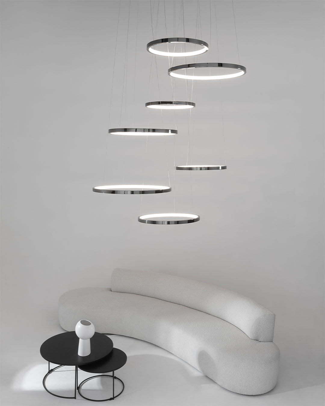 Lámpara de techo LED moderna con aros metálicos sobre sofá curvo blanco y mesa negra.