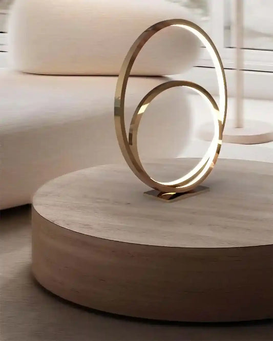 Lámpara de mesa moderna dorada con diseño circular sobre mesa de madera en sala minimalista