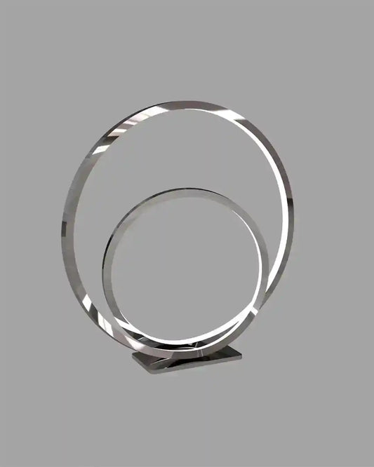 Lámpara de mesa LED moderna en forma de aro doble color plateado sobre fondo gris.