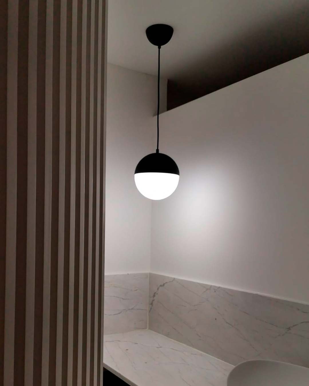 Lámpara colgante negra moderna iluminando baño con paredes y lavamanos de mármol blanco