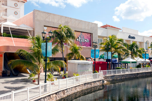 Centro comercial moderno junto a un canal, con palmeras y tiendas al fondo bajo cielo soleado.