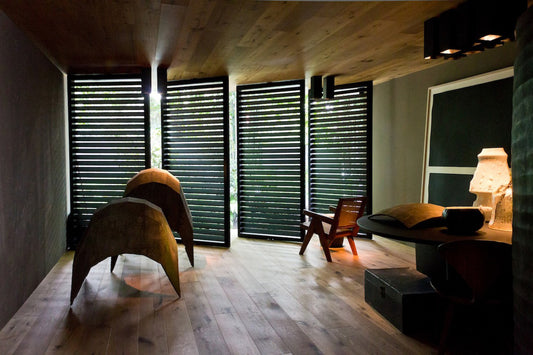 Habitación moderna con piso de madera, sillas de diseño y luz natural filtrada por persianas oscuras.